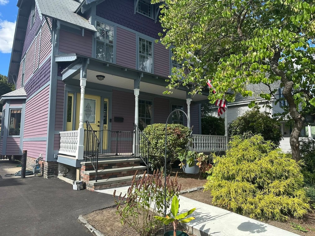 This is an image from Power Painting Plus, located in Massachusetts. It shows a freshly painted Victorian-style home with purple siding, blue-gray trim, and a yellow front door, highlighting the company’s detailed exterior painting services.