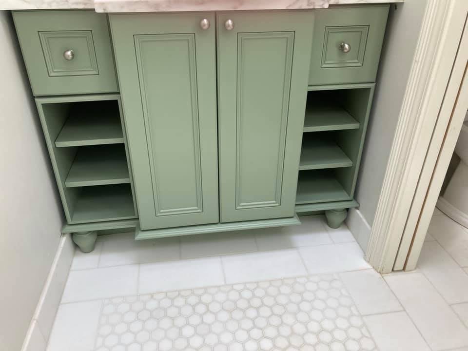 This is an image from JS Professional Painters Inc company, located in Glencoe, IL The image is a custom-painted sage green bathroom vanity with open shelving, drawers, and white marble countertop on tiled floor