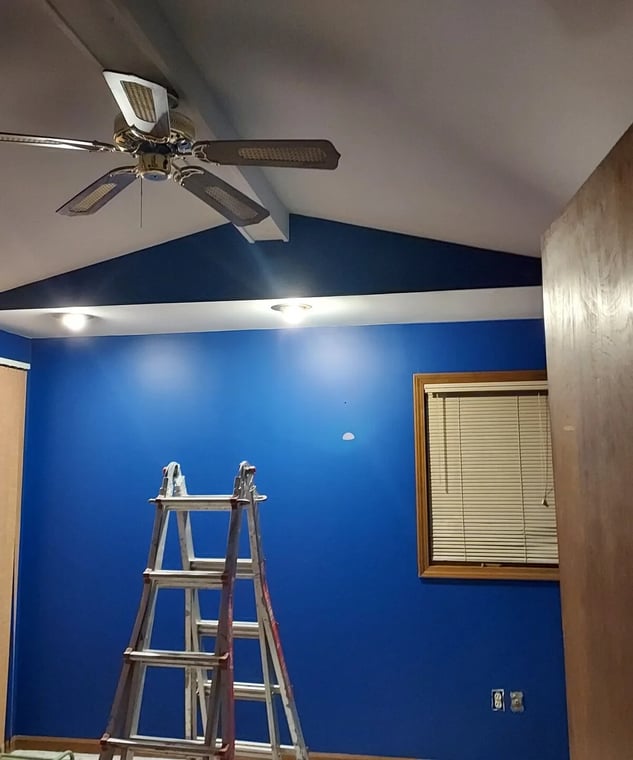 This is an image from Pastels Painting CO company, located in Warren, MI The image is an interior painting of a room with a bold blue accent wall, vaulted ceiling, and ceiling fan, featuring a ladder setup for painting near a wood-framed window.