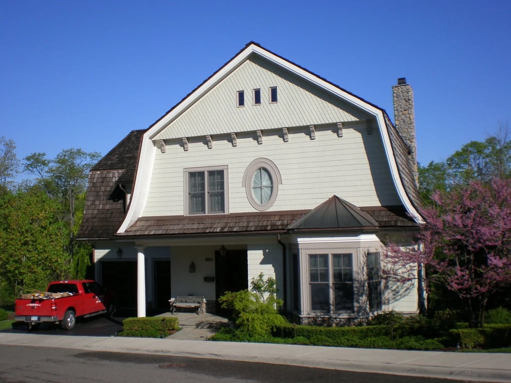 This is an image from The Exterior Painting Experts - F&H Painting company, located in Oxford, MI The image is a beautiful two-story home with fresh exterior paint in neutral tones, featuring unique roof lines, detailed trim work, and a red pickup truck parked in the driveway.