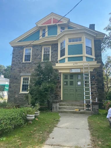 This is an image from Eli Painting, located in Massachusetts. It shows a stone exterior house with green doors and yellow trim, where a ladder is set up for exterior painting work.