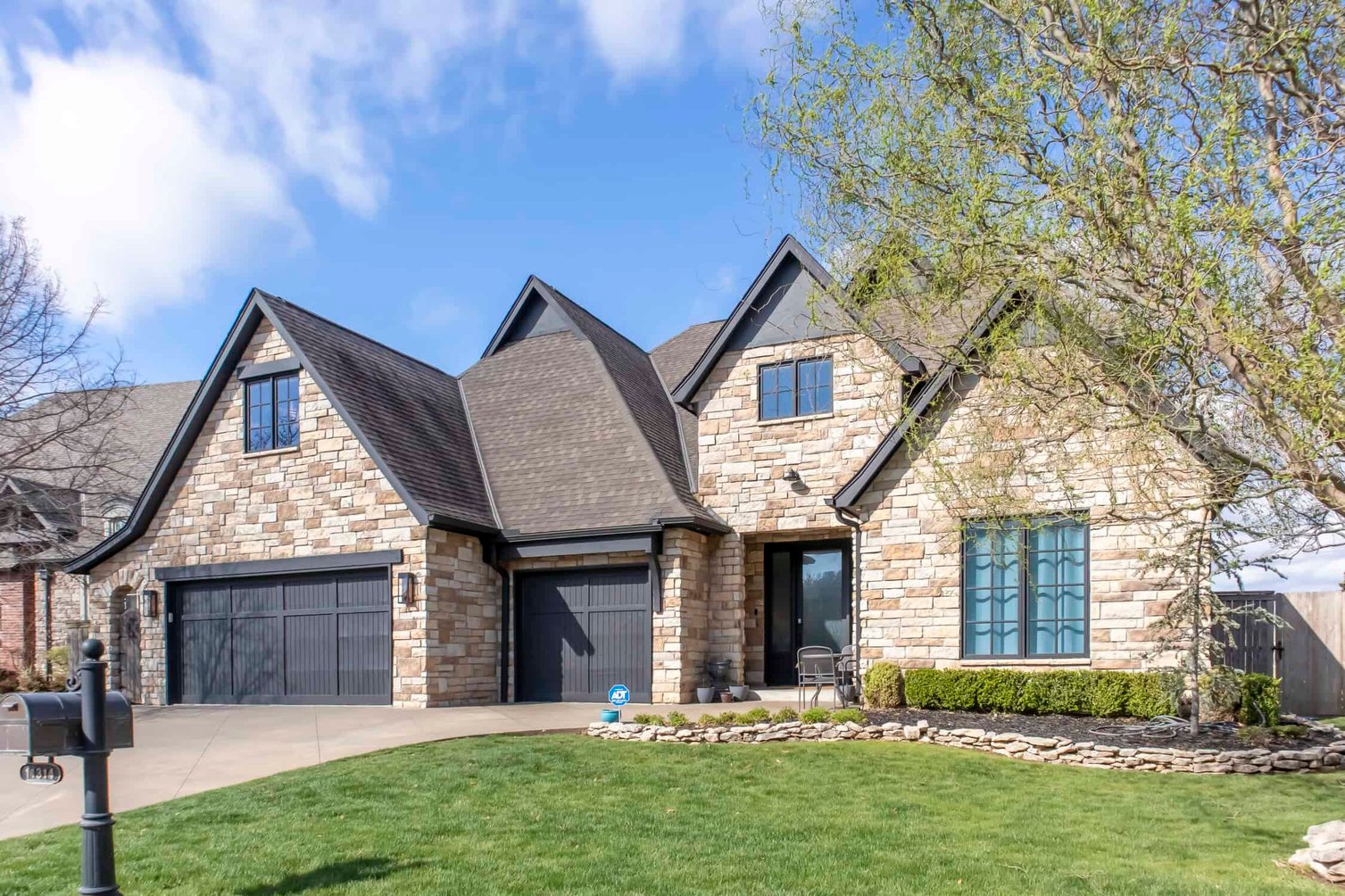 This is an image from Impressions Painting Company, located in Oklahoma. The photo shows a brick-front house with a light gray garage door, landscaped shrubs, and a spacious driveway.