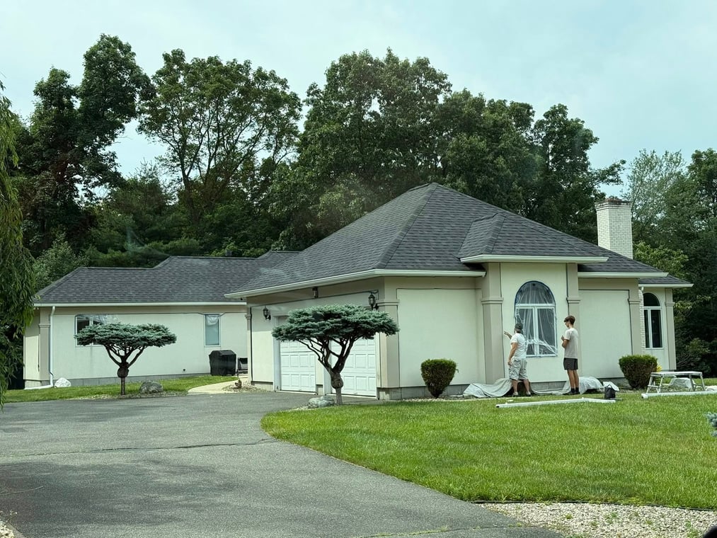 Two professional painters are working on the front exterior of a light beige stucco house. They have covered a large arched window with protective plastic and laid down white drop cloths on the ground as they prepare to paint the wall, indicating an exterior painting project in progress.
