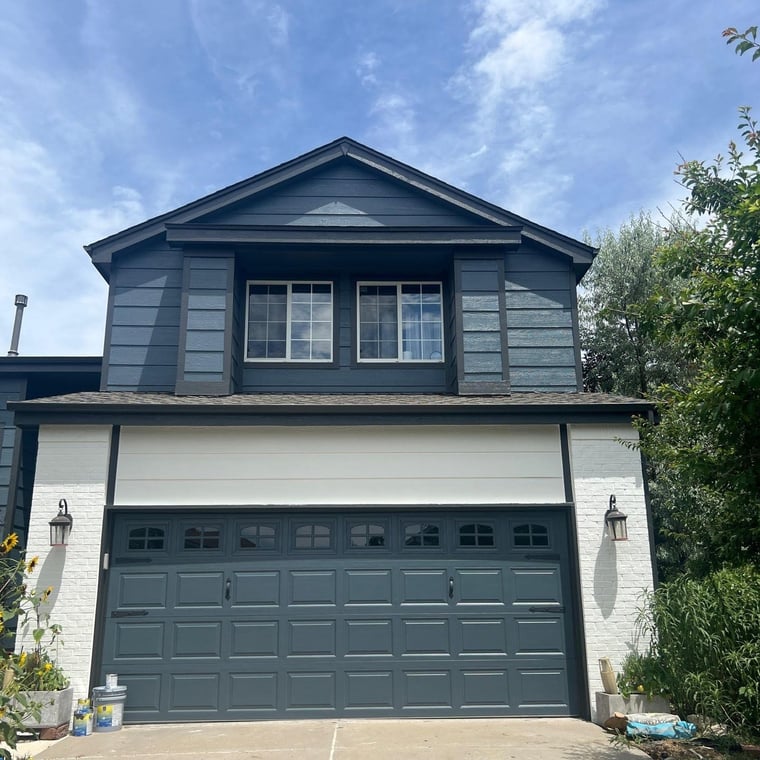 This is an image from Paint Denver company, located in Denver, CO The image is a two-story house with fresh exterior paint in dark slate blue and white, featuring a modern garage door and crisp trim by Paint Denver