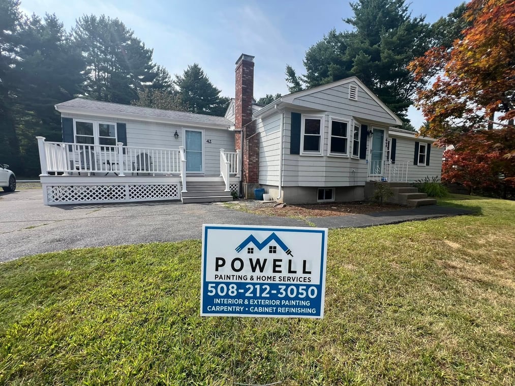 This is an image from Powell Painting & Home Services, located in Massachusetts. The photo shows a freshly painted light gray home with white trim, a blue front door, and a Powell Painting yard sign displayed in the front lawn