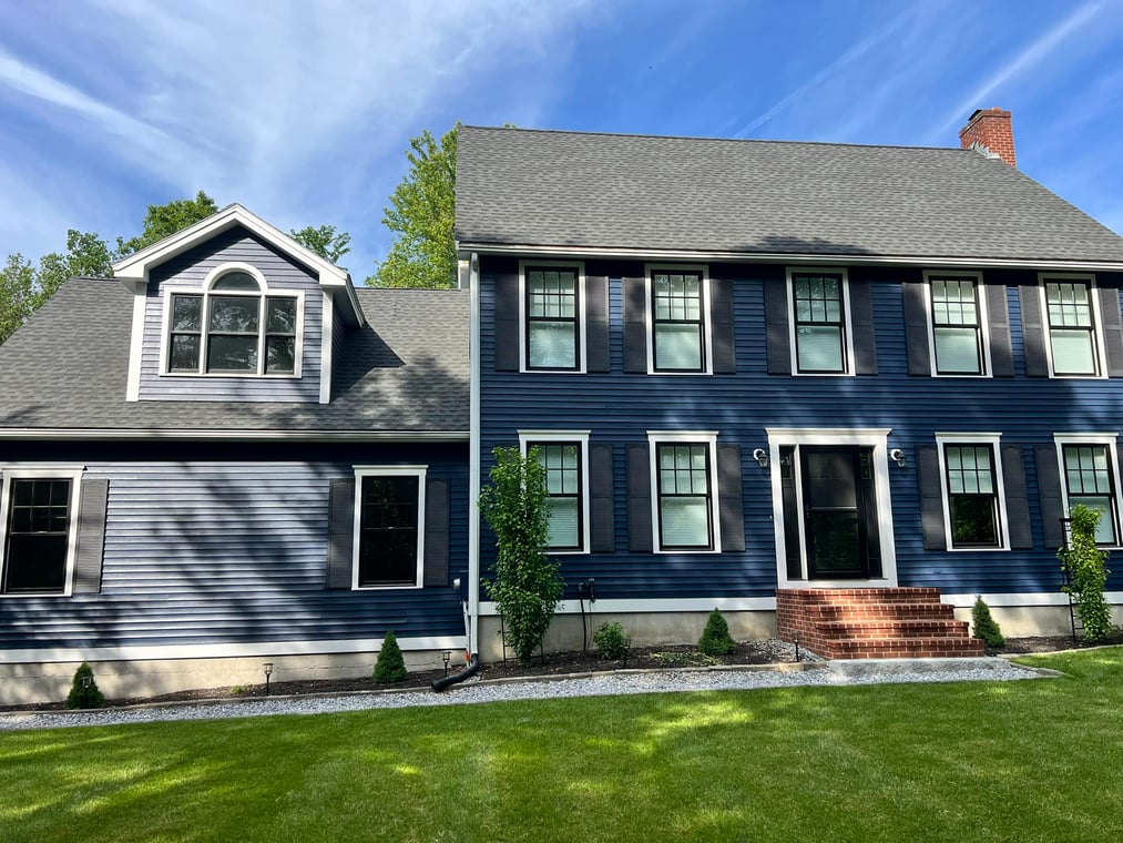 This is an image from Amazing Painting Company, LLC, located in Massachusetts. The photo shows a large colonial-style home with dark blue siding, white trim, and black shutters, freshly painted for a clean and updated look.
