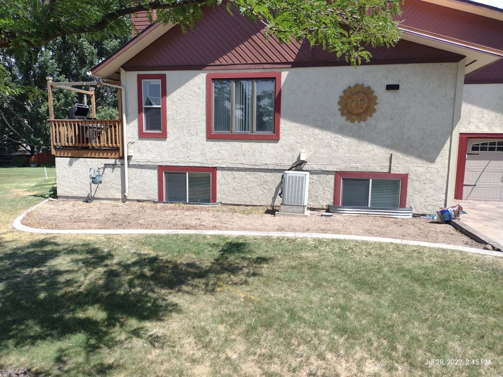 This is an image from Horizon Painting LLC company, located inPocatello, ID The image is a side view of a two-story beige stucco home with red trim, small deck, basement windows, and landscaped yard in Pocatello, Idaho.