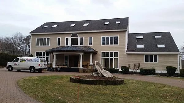This is an image from Costa Painting Service MA, located in Massachusetts. It shows a large beige two-story home with multiple roof windows, a front porch, and a Costa Painting Service van parked in the driveway.