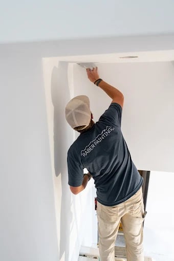This is an image from Raber Painting company, located in Gunnison, CO The image is a Raber Painting crew member in branded shirt prepping or painting a white interior ceiling corner using a step ladder