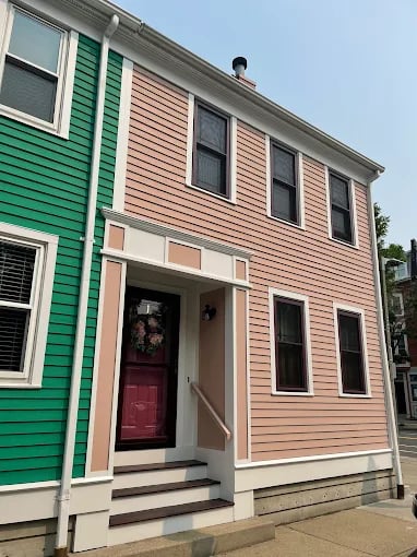 This is an image from CertaPro Painters of Boston. It shows the exterior of a two-story home painted in soft pink with white trim and a red front door.