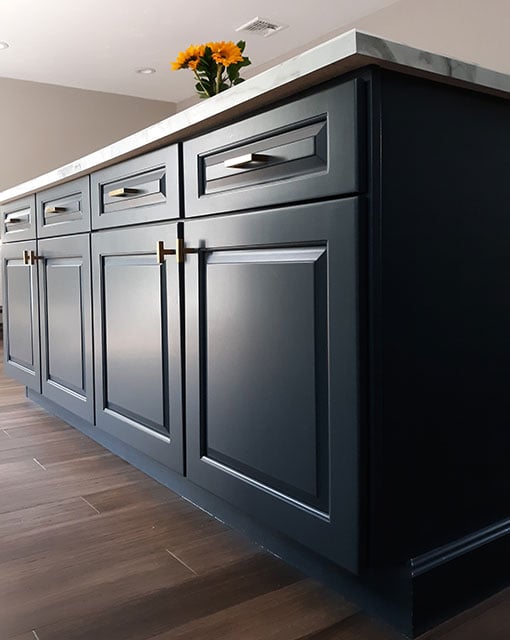 This is an image from Linda Loo Designs Cabinet Refinishing & Refacing, showing a modern kitchen island with dark blue cabinetry, marble countertop, and brass handles. A small vase with bright sunflowers decorates the counter.