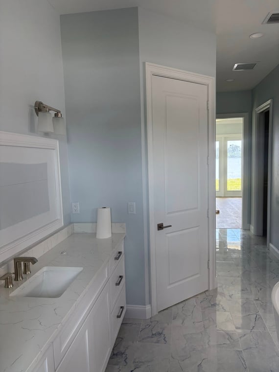 This is an image from Coastline Painting Professionals company, located in Grant-Valkaria, FL. The image is a modern bathroom with light blue painted walls, white vanity with marble countertop, and glossy marble tile flooring leading to a sunlit room