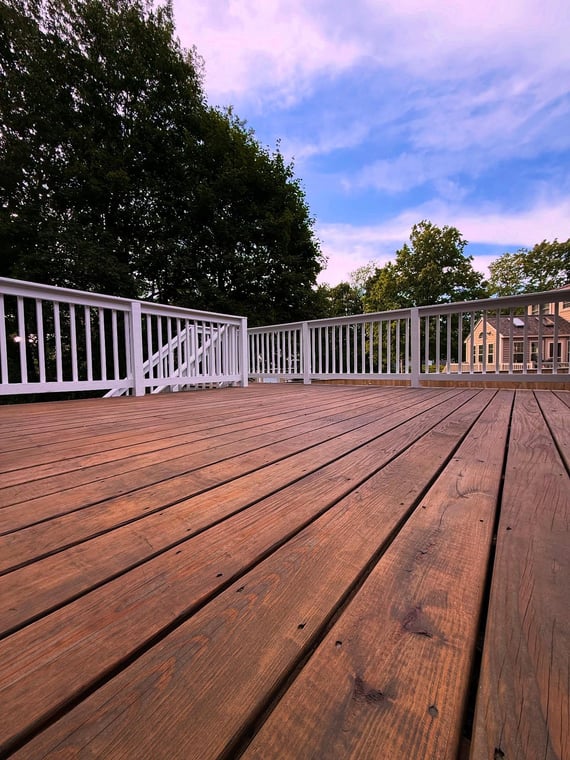A freshly stained wooden deck with clean white railings, showcasing a natural finish against a backdrop of trees and a blue sky.