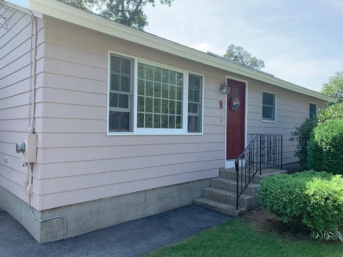 This is an image from Castle Complements Painting Co., located in Massachusetts. It shows the exterior of a single-story home painted in light beige with a red front door and white trim.