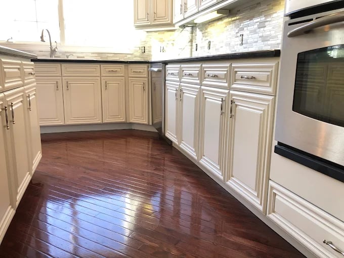 This is an image from TotalPro Painting & Remodeling, showcasing a kitchen with newly refinished cabinetry. The cabinets are painted in a crisp cream tone with detailed molding, paired with polished wooden floors that reflect light beautifully.