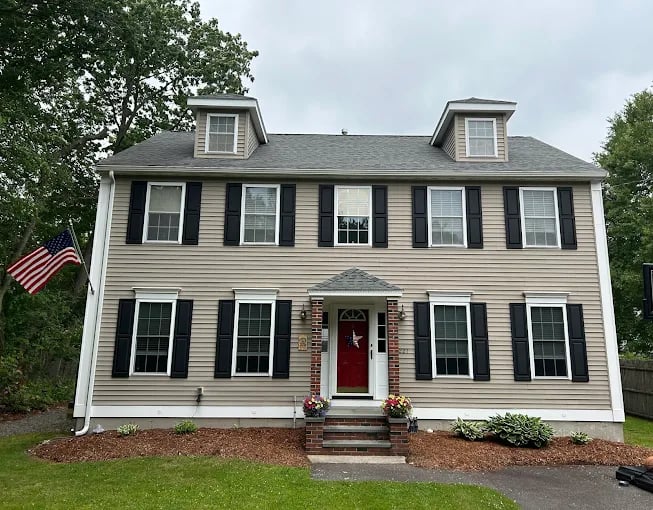 This is an image from CertaPro Painters of Needham, located in Massachusetts. The photo shows a freshly painted beige two-story colonial-style home with black shutters, a bright red front door, and neatly landscaped surroundings