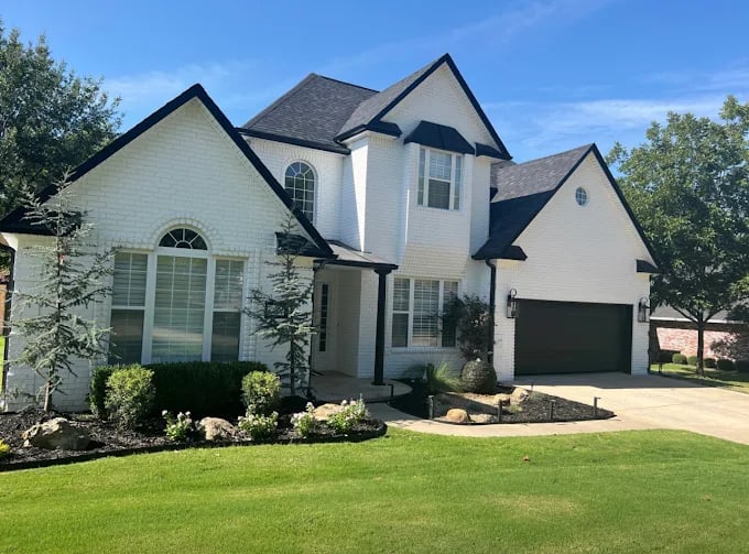 This is an image from Derheim Painting, located in Oklahoma. The photo shows a freshly painted two-story white brick home with black trim, dark roofing, and a neatly landscaped front yard.
