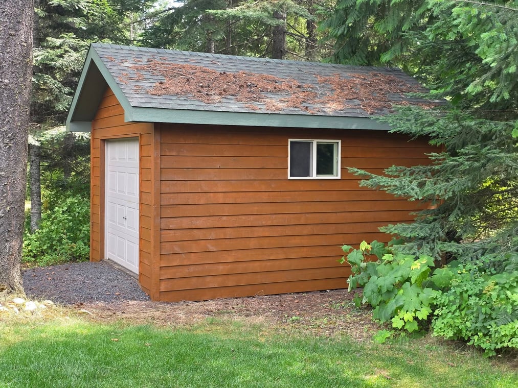 This is an image from Honey Do Painting LLC company, located in Bonners Ferry, ID The image is a freshly stained wood storage shed with green-trimmed roof, single window, and white roll-up door, surrounded by forest trees and landscaped greenery, project by Honey Do Painting
