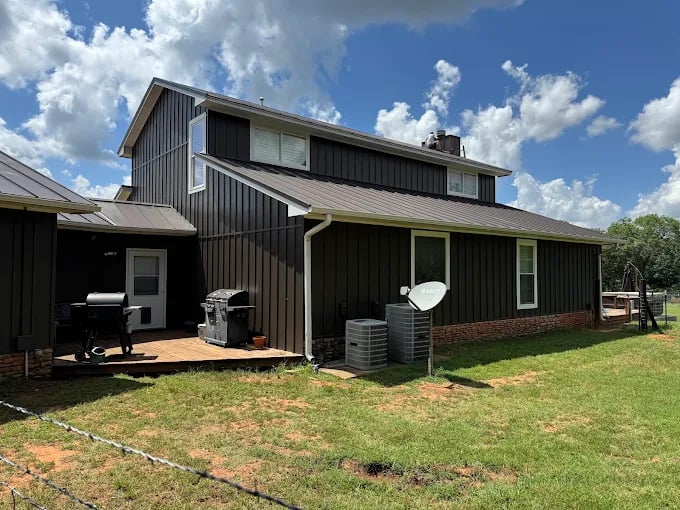 This is an image from Fresh Coat Painters of Oklahoma City, located in Oklahoma. The photo shows a two-story home with freshly painted dark brown board-and-batten siding, white trim, and a metal roof under a bright blue sky.