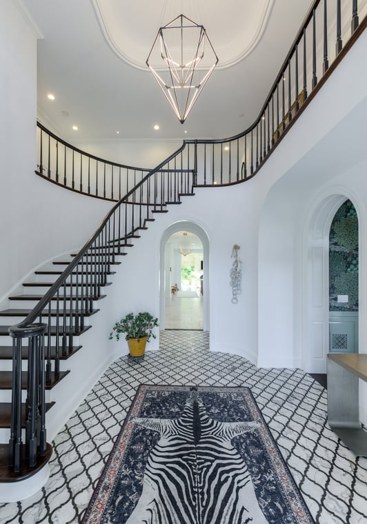 This is an image from Maurer Painting LLC Company, located in Boulder, CO The image is a freshly painted luxury foyer with white walls, grand curved staircase with dark wood railing, modern chandelier, and patterned tile floor by Maurer Painting in Colorado. .