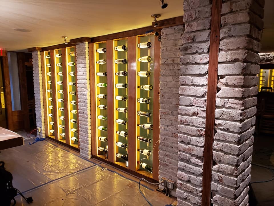 This is an image from https://specializedfinishes.com/ company, located inGrand Junction, CO The image is a custom wine cellar wall with illuminated bottle display and exposed brick columns by Specialized Finishes