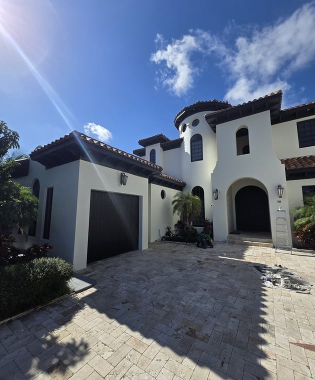 This is an image from Big Family Painting Co  company, located in Boca Raton, FL. The image is a luxury mediterranean-style home exterior painted white with dark trim and clay roof tiles, featuring paver driveway and painting tools visible