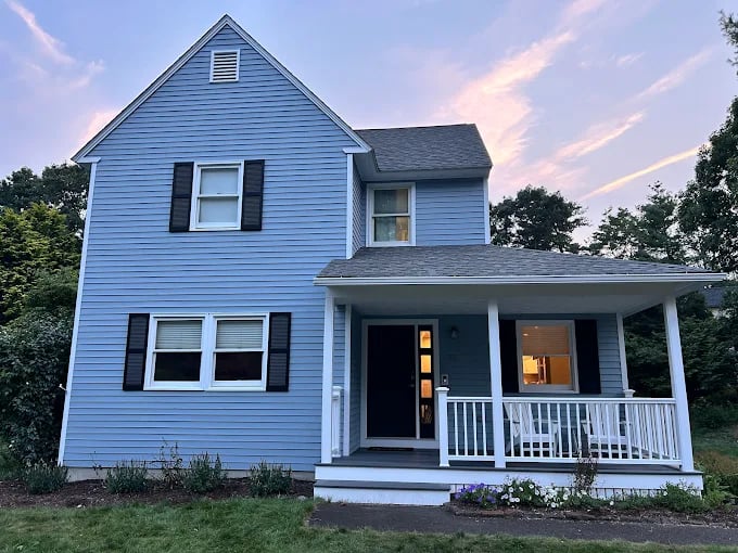 This is an image from Aurora Exterior Painting Inc., located in Massachusetts. The photo shows a two-story home with freshly painted light blue siding, white trim, and black shutters, giving it a clean and modern appearance.