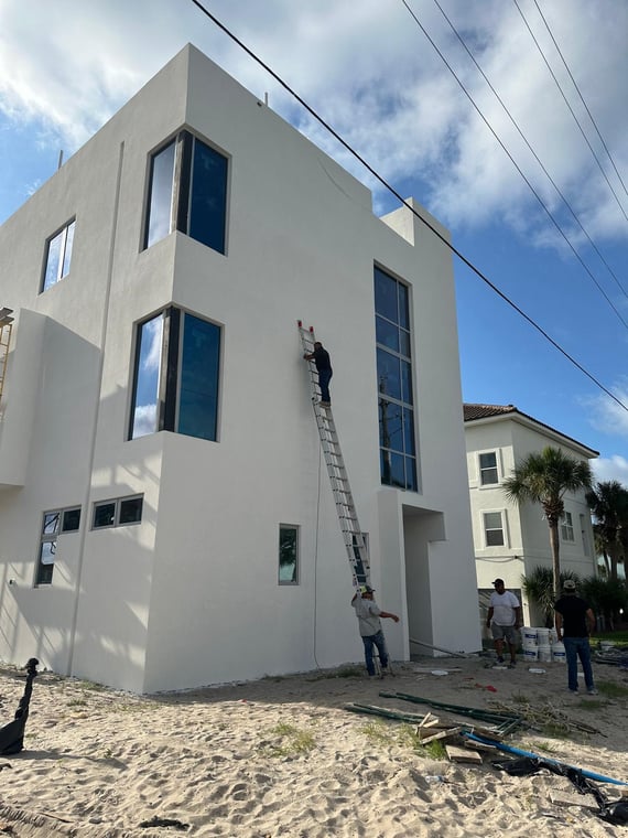 This is an image from Falcon Remodeling Express LLC company, located in Fort Walton Beach, FL. The image is an exterior painting crew working on a modern beachfront home using ladders and rollers for a smooth white stucco finis