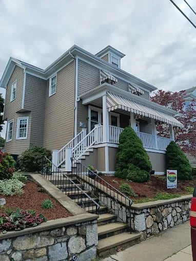 This is an image from Meticulous Painting, located in Massachusetts. The photo shows a freshly painted two-story beige home with white trim, decorative awnings, and neatly landscaped shrubs along a stone retaining wall.