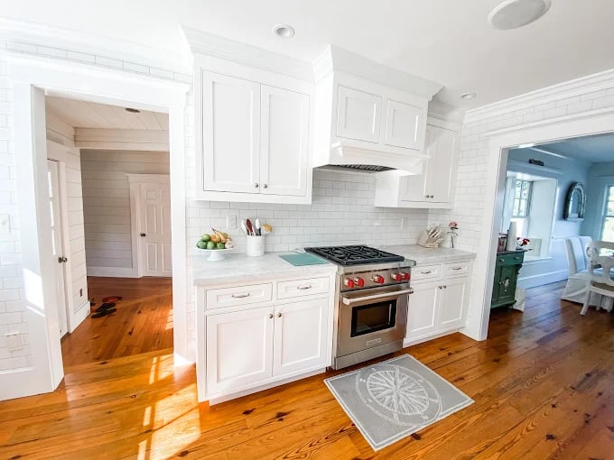 This is an image from Stewart Painting, located in Massachusetts. The photo shows a bright kitchen with freshly painted white cabinets, subway tile backsplash, and natural wood flooring.