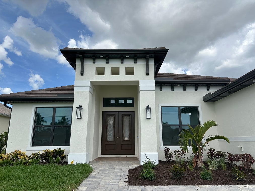 This is an image from Cook Homes company, located in Cape Coral, FL. The image is a modern single-story white stucco home with dark trim and landscaped front entrance built by Cook Homes