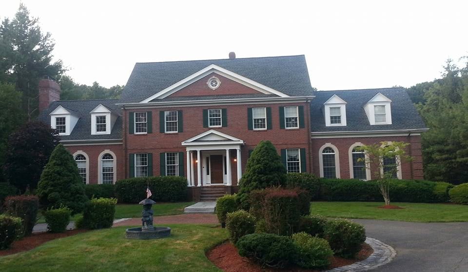 This is an image from Allan Painting, located in Massachusetts. The photo shows a stately red brick colonial-style home with green shutters, white trim, and a manicured lawn surrounded by lush greenery.