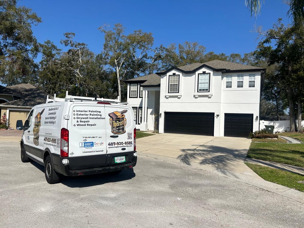 This is an image from TopBrush Painting company, located in Altamonte Springs, FL. The image is a TopBrush Painting service van parked in front of a newly painted two-story white home with black trim, showcasing residential exterior painting in Florida