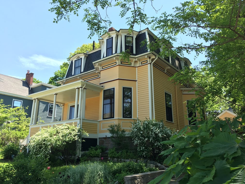 This is an image from Catchlight Painting, located in Massachusetts. It shows a yellow Victorian-style home with black roof shingles and white trim, surrounded by greenery and trees.