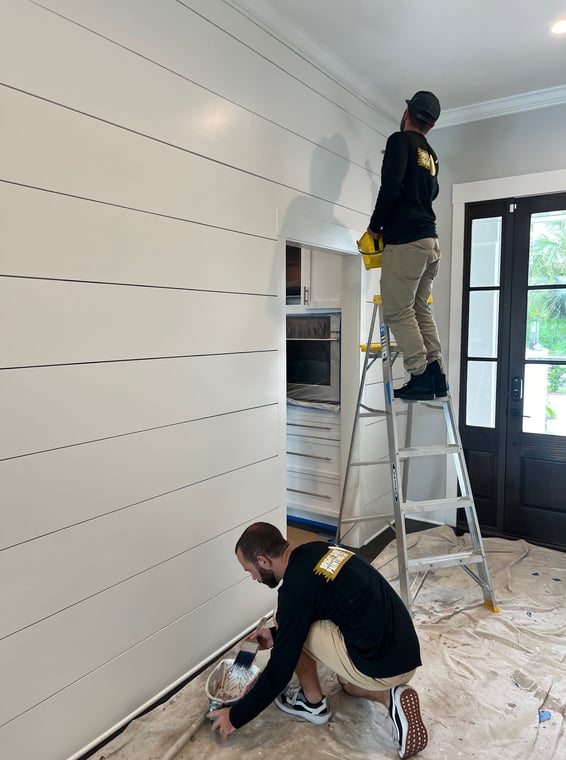 This is an image from Bright Side Painting Of Jax company, located in Jacksonville Beach, FL. The image is an interior house painters from Bright Side Painting applying fresh white paint to shiplap wall in modern home using brush and ladder