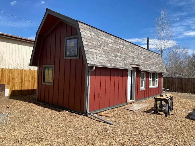 This is an image from Flawless Painting LLC company, located in Longmont, CO The image is a small red barn-style shed with brown shingle roof, white trim, and multiple windows, situated in a fenced backyard under a clear blue sky