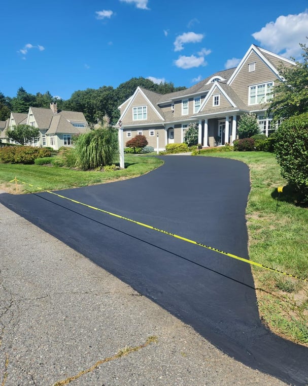 This is an image from Right Track Painting & Sealcoating, showing a freshly sealcoated driveway in front of a large tan house. The surface has a smooth black finish with caution tape placed across the entrance.