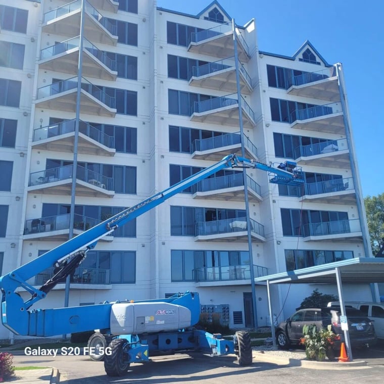 This is an image from America's Commercial Painting company, located in Troy, MI The image is a commercial exterior painting project on a multi-story condominium building with balconies, featuring a boom lift used by painters to access high areas for facade painting and maintenance.