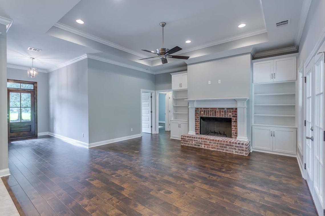 This is an image from Schmidt Painting, located in Massachusetts. It highlights an interior living room painted in a light gray tone by Schmidt Painting, with white trim, built-in shelving, and a red brick fireplace. The space features dark wood flooring and a ceiling fan.