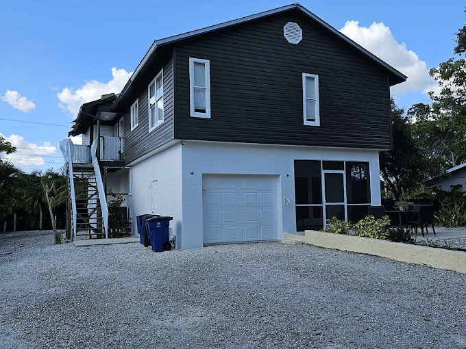 This is an image from Prime Design Painting company, located in Naples, FL. The image is a two-story house with freshly painted black upper siding and white lower exterior by Prime Design Painting, showing a garage, gravel driveway, and outdoor staircase. 