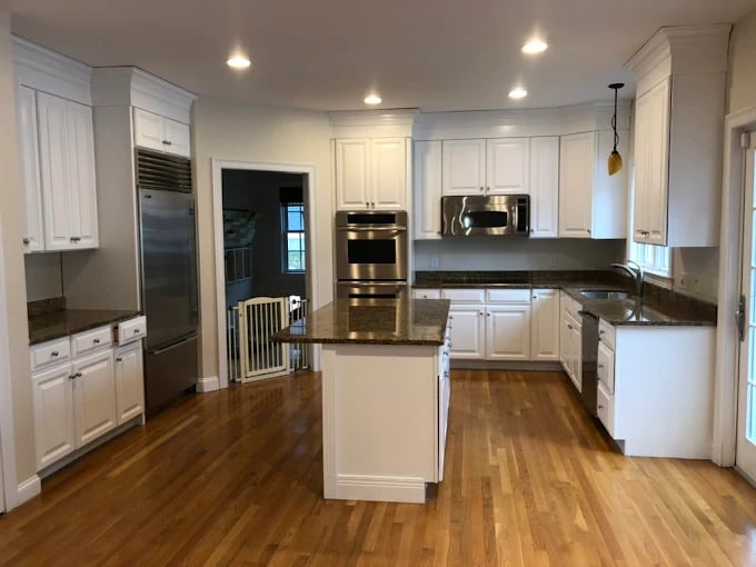 This is an image from JK Painting Service, located in Massachusetts. The photo shows a modern kitchen with freshly painted white cabinets, stainless steel appliances, granite countertops, and polished hardwood flooring.