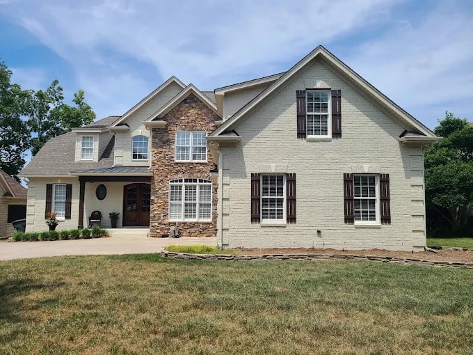 This is an image from Five Star Painting, showing a large two-story home with stone and brick accents. The exterior is painted a light beige with dark brown shutters, highlighting the contrast between natural textures and fresh paint.