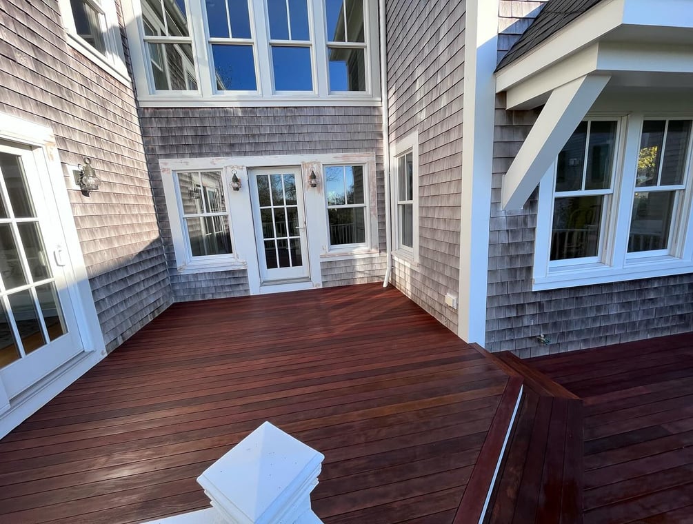 This is an image from Cape Cod Fine Finishes LLC, located in Massachusetts. It shows a newly refinished wooden deck with rich dark tones, set against a shingle-style home with multiple white-trimmed windows and glass doors.