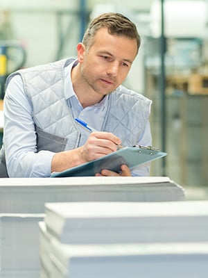 A man taking notes on a clipboard while leaning over stacks of paper.