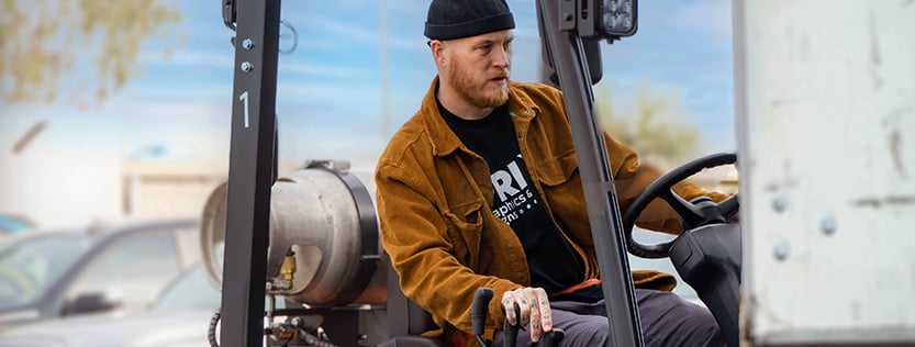 man wearing a black beanie and brown jacket operating a forklift.