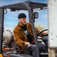 man with a black beanie and brown jacket driving a forklift.