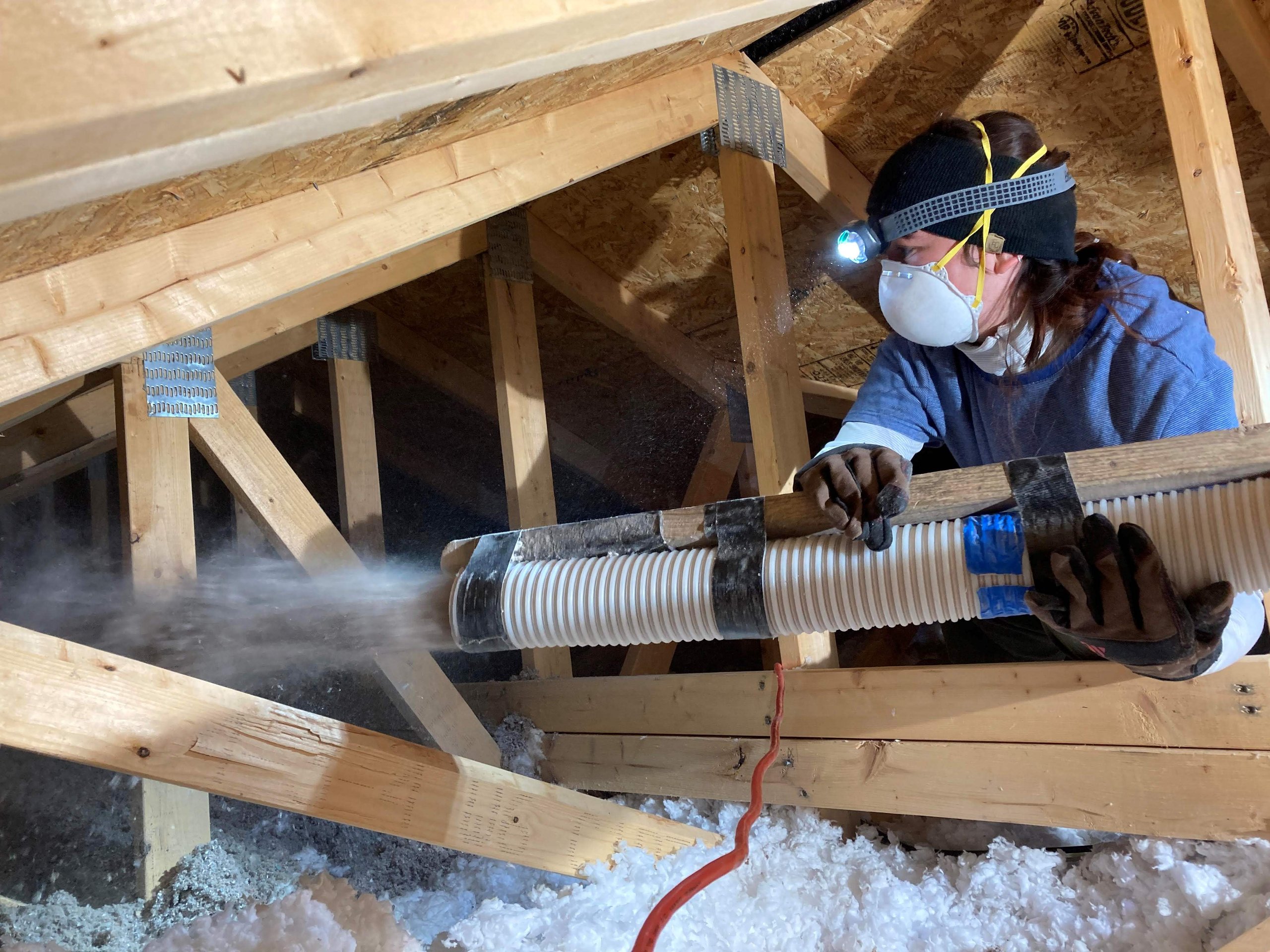 A student wearing PPE and a headlamp working on filling a rafter space on campus with insulation.