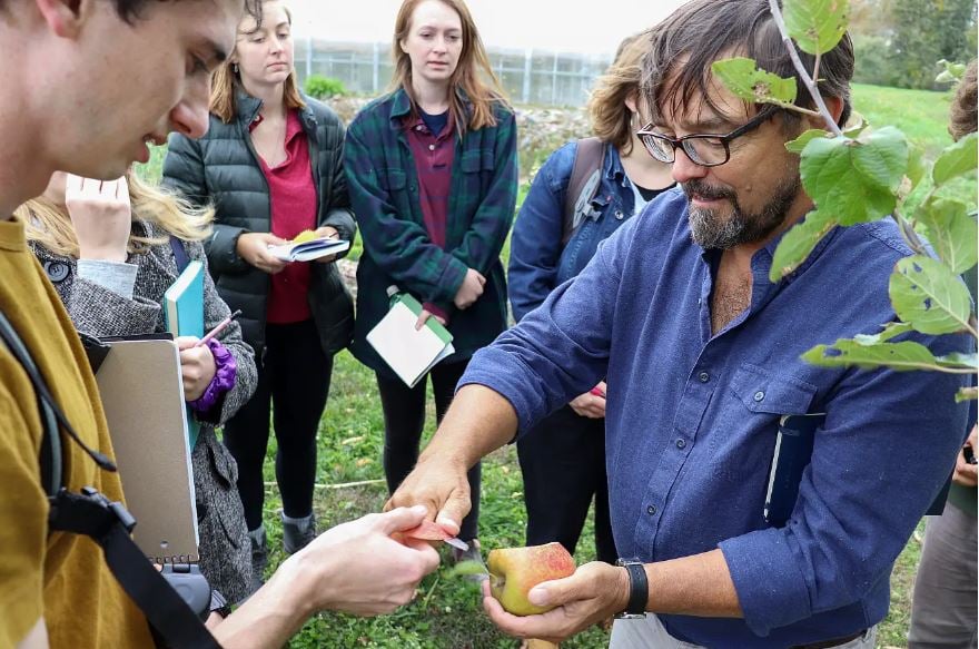 professor Todd Little-Siebold with a class outdoors, handing freshly-cut apple slice to a student