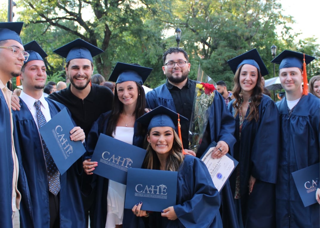 Graduates in navy gowns holding CAHE diplomas outdoors