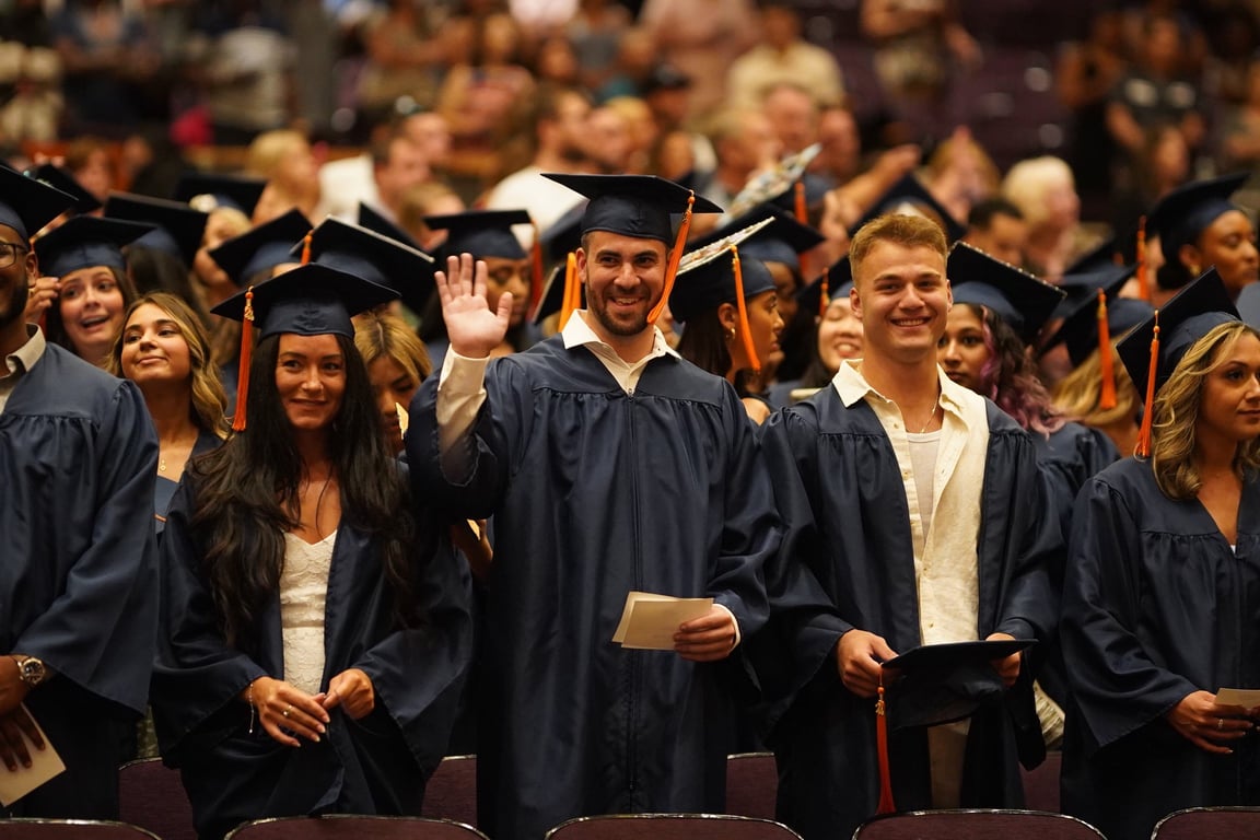 CAHE graduates smiling outdoors in navy blue regalia with bright orange tassels.